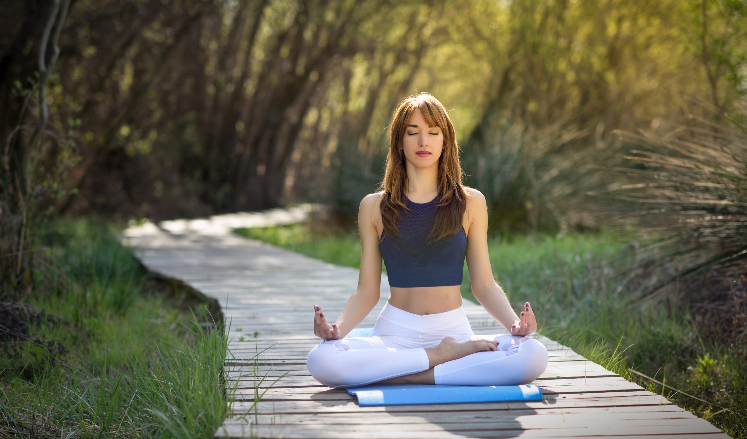 Young woman doing yoga in nature. Female wearing sport clothes in lotus figure. Girl sitting on wooden road.